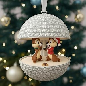 Op de afbeelding: Een kerstversiering met Chip en Dale in een koepelvormig ontwerp. Eén eekhoorn draagt een kerstmuts en houdt een ringdoos vast, terwijl de andere een groene sjaal draagt. De versiering heeft een wit lint om op te hangen.