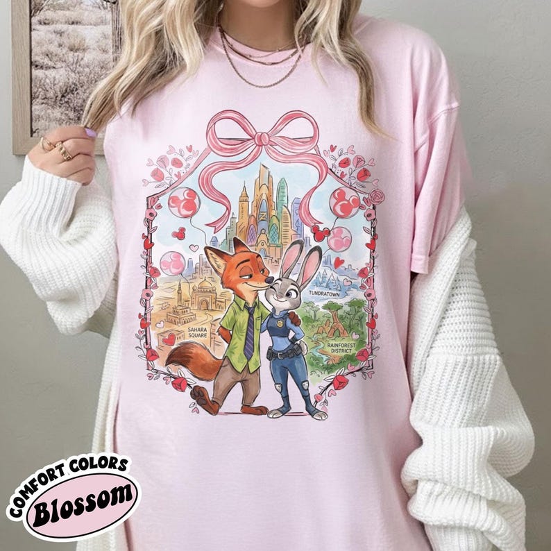 May include: Pink t-shirt with a graphic of Nick Wilde and Judy Hopps from Zootopia, framed by a pink bow and floral accents. The shirt features the text "COMFORT COLORS Blossom".