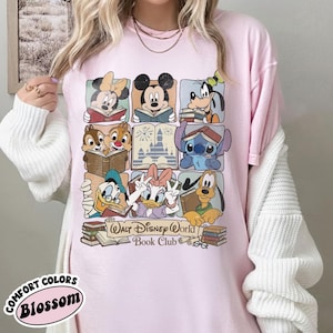 Puede incluir: Camiseta rosa claro con un gr&aacute;fico colorido de personajes de Disney leyendo libros, incluyendo Mickey Mouse, Minnie Mouse y Goofy. El texto "Walt Disney World Book Club" est&aacute; impreso debajo de los personajes. La camiseta tambi&eacute;n tiene el texto "Comfort Colors Blossom".
