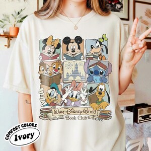 Puede incluir: Camiseta de color marfil con un gr&aacute;fico colorido de personajes de Disney leyendo libros. El dise&ntilde;o incluye Mickey Mouse, Minnie Mouse, Goofy, Donald Duck, Daisy Duck, Chip y Chop, Stitch y Pluto. El texto dice "Walt Disney World Book Club."