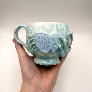 May include: A handmade ceramic mug in shades of blue and cream, featuring a sculpted fish and seaweed design. The mug has a rounded base, a sturdy handle, and is approximately 10 cm tall.