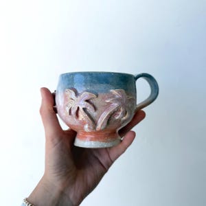 May include: A ceramic mug with a blue rim, a pink and orange body, and a white base. The mug features two palm tree designs carved into the body. The mug has a handle and is held by a person.