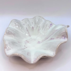 May include: A decorative ceramic bowl with a scalloped edge. The bowl has a pearlescent finish with shades of white, light blue, and hints of purple. The rim of the bowl has a light brown colour.