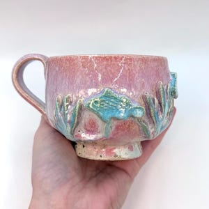 May include: A handcrafted ceramic mug with a pink and blue glaze. The mug has a sculpted design of fish and seaweed around the base. The handle is curved, and the mug is held in a hand.