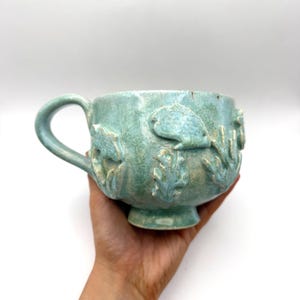 May include: A handmade, seafoam green ceramic mug with a large handle. The mug features embossed aquatic designs, including fish and seaweed. The mug is held in a hand against a white background. The mug has a footed base.