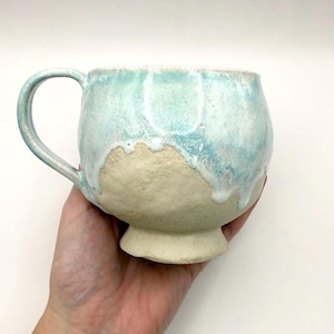 May include: Handmade ceramic mug with a rounded body and a footed base. The mug has a light blue glaze that drips over natural, unglazed beige clay. The handle is light blue. A unique, handcrafted piece for enjoying beverages.