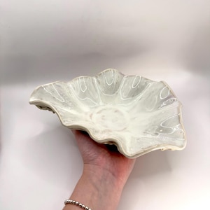 May include: A shallow, scalloped ceramic bowl with a glossy, off-white glaze featuring subtle, swirling grey marbling throughout the interior surface. The bowl has a wide, irregular, wavy rim that mimics the organic shape of a flower petal.