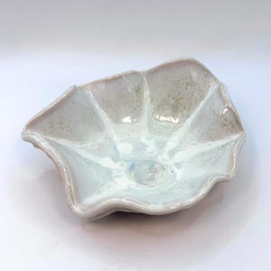 May include: A ceramic bowl with an unusual, organic form. The bowl has a light, speckled glaze with hints of blue and white. The edges have a subtle, wavy design, enhancing its artistic appeal. This decorative bowl is suitable for serving or display.