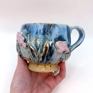 May include: A handcrafted ceramic mug with a blue and gold glaze. The mug has pink floral decorations and a speckled tan base. The handle is blue. This unique mug is suitable for tea or coffee.