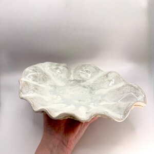May include: A shallow, round ceramic bowl with a wavy, scalloped edge. The surface features a glossy, off-white glaze with subtle, mottled grey and beige speckling throughout, creating a textured appearance across the entire interior basin.