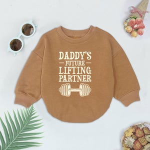 Może przedstawiać: Brązowa bluza z nadrukiem "Daddy's Future Lifting Partner" i grafiką hantle.