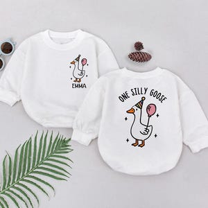 以下が含まれることがあります： 長袖の白いベビーロンパース2枚。1つはパーティーハットと風船を持った漫画のガチョウで、下に「EMMA」という名前があります。もう1つは、同様のガチョウのデザインで「ONE SILLY GOOSE」と書かれています。