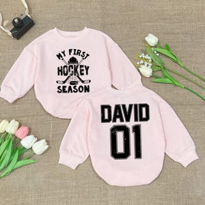Może przedstawiać: Dwie r&oacute;żowe bluzy z czarnym nadrukiem. Jedna bluza ma napis "My First Hockey Season" z grafiką hokejowej kija i krążka. Druga bluza ma napis "David 01".