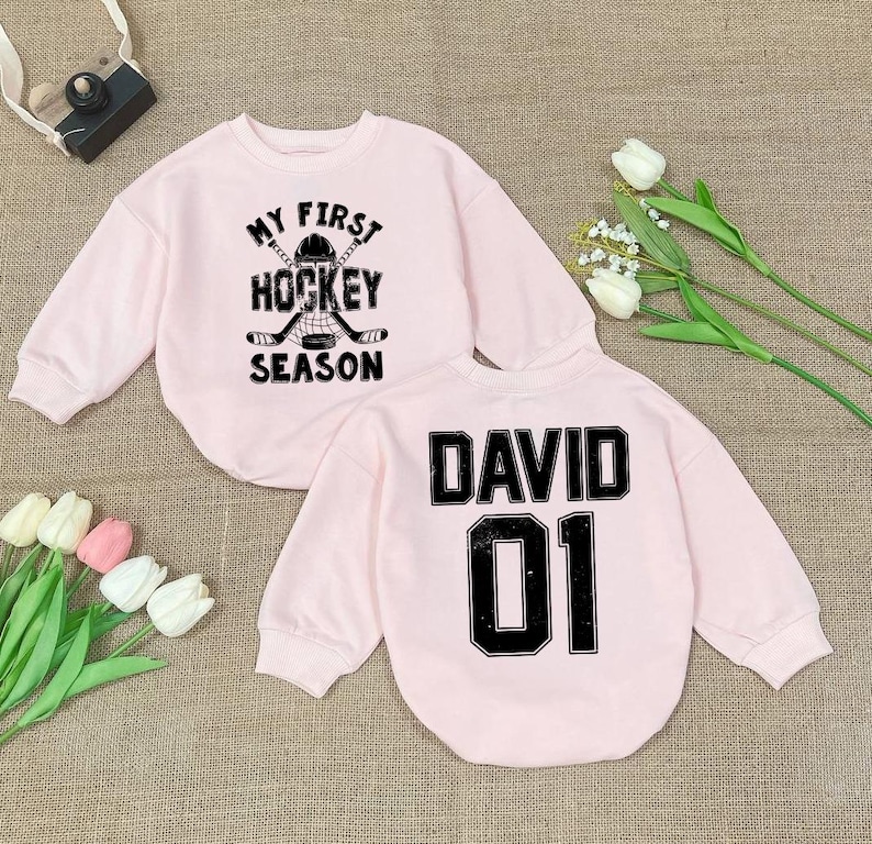 Może przedstawiać: Dwie r&oacute;żowe bluzy z czarnym nadrukiem. Jedna bluza ma napis "My First Hockey Season" z grafiką hokejowej kija i krążka. Druga bluza ma napis "David 01".
