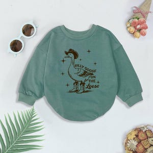 May include: A light green sweatshirt with a graphic of a goose wearing a cowboy hat and boots. The text "Silly Goose on the Loose" is printed on the sweatshirt.