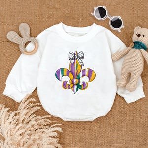 May include: White long-sleeve baby onesie with a purple, green, and gold fleur-de-lis design and a white bow with black polka dots.