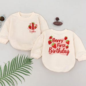 May include: Two cream-colored long-sleeved shirts with a strawberry theme. One shirt says "Berry First Birthday" with strawberries around the text. The other shirt has a number one and the name "Sophia" printed on it.