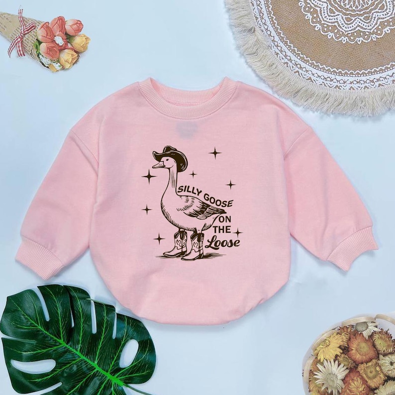 May include: A pink long-sleeved sweatshirt with a graphic of a goose wearing a cowboy hat and boots. The text "Silly Goose on the Loose" is printed below the goose.
