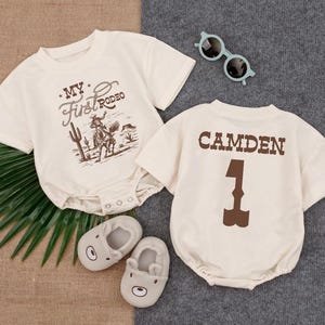 May include: Two cream-colored short-sleeve baby bodysuits. One features a brown graphic of a cowboy on a horse with the text "MY First RODEO". The other displays the name "CAMDEN" above the number "1" in brown. Includes light blue sunglasses.