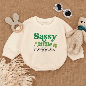 May include: A cream colored long-sleeved onesie with the text "Sassy little lassie" in green and black lettering. There are two green shamrock designs on the onesie.