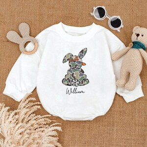 May include: White long-sleeve baby onesie with a camouflage bunny wearing a backwards baseball cap. The bunny is holding a carrot and the onesie is personalized with the name "William".