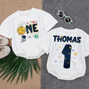Puede incluir: Dos bodies blancos para bebés con gráficos de temática espacial. Uno presenta la palabra "ONE" con planetas y un astronauta. El otro tiene el nombre "THOMAS" y el número "1" con estrellas. Incluye zapatos de bebé y gafas de sol.