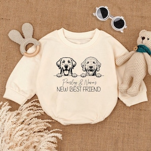 May include: A cream-colored baby onesie with black illustrations of two dogs and the text "Paisley & Nova's New Best Friend".