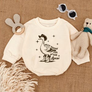 May include: A cream-colored baby onesie with a black and white illustration of a goose wearing a cowboy hat and boots. The text "Silly Goose on the Loose" is printed below the goose.
