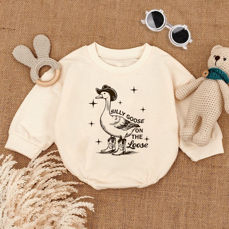 May include: A cream-colored baby onesie with a black and white illustration of a goose wearing a cowboy hat and boots. The text "Silly Goose on the Loose" is printed below the goose.