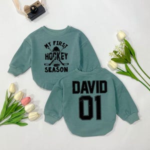 Może przedstawiać: Szarfa w kolorze turkusowym z napisem "My First Hockey Season" i grafiką hokejowej kija i krążka na przodzie. Z tyłu szarfy znajduje się imię "David" i numer "01" w kolorze czarnym.