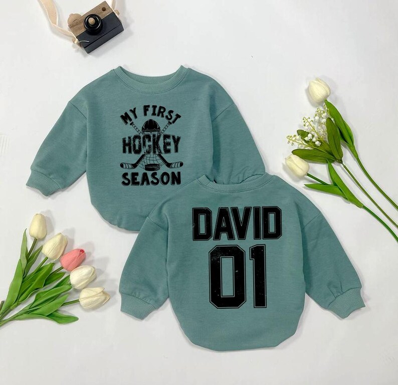 Może przedstawiać: Szarfa w kolorze turkusowym z napisem "My First Hockey Season" i grafiką hokejowej kija i krążka na przodzie. Z tyłu szarfy znajduje się imię "David" i numer "01" w kolorze czarnym.