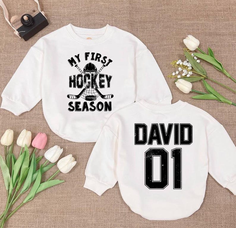 Może przedstawiać: Dwie białe koszulki z długimi rękawami. Jedna koszulka ma napis "My First Hockey Season" z grafiką hokejowej kija i krążka. Druga koszulka ma napis "David 01".