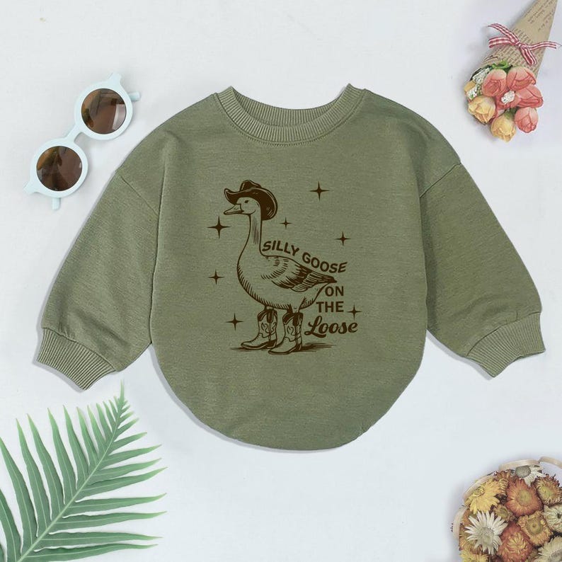 May include: A green long-sleeved sweatshirt with a brown graphic of a goose wearing a cowboy hat and boots. The text "Silly Goose on the Loose" is printed below the goose.