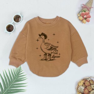 May include: A brown long-sleeved sweatshirt with a graphic of a goose wearing a cowboy hat and boots. The text "Silly Goose on the Loose" is printed on the sweatshirt.