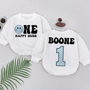 May include: Two white long-sleeved shirts with black text. One shirt says "ONE HAPPY DUDE" with a smiley face. The other shirt says "BOONE" with a number "1" in a blue and white checkered pattern.