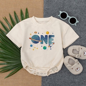 May include: Cream baby romper featuring a space-themed design with the word "ONE", planets, stars, and a rocket. The romper has short sleeves and popper fastenings. The image also includes bear-shaped baby shoes and sunglasses.