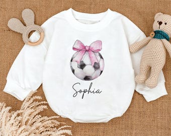 May include: White baby bodysuit with long sleeves. The front features a watercolor soccer ball design with a pink bow and the name "Sophia" in script. A wooden bunny-shaped teether and a stuffed teddy bear are in the background.