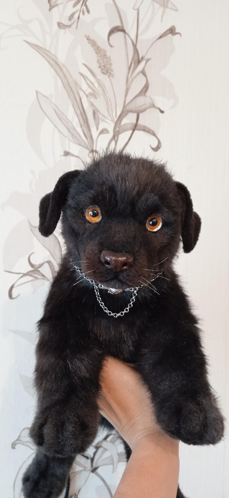 May include: A black plush puppy with amber eyes and a silver chain collar. The soft toy has dark fur and is held up in front of a patterned background. The puppy's expression is alert and curious.