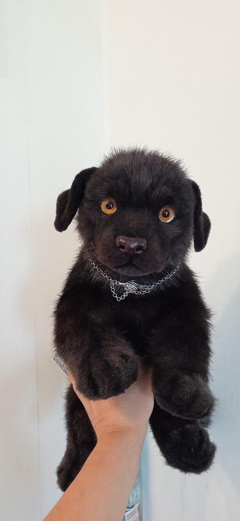 May include: A black plush puppy with amber eyes and a silver chain collar. The soft toy has a furry texture and is held up against a white background. The puppy's nose is brown.