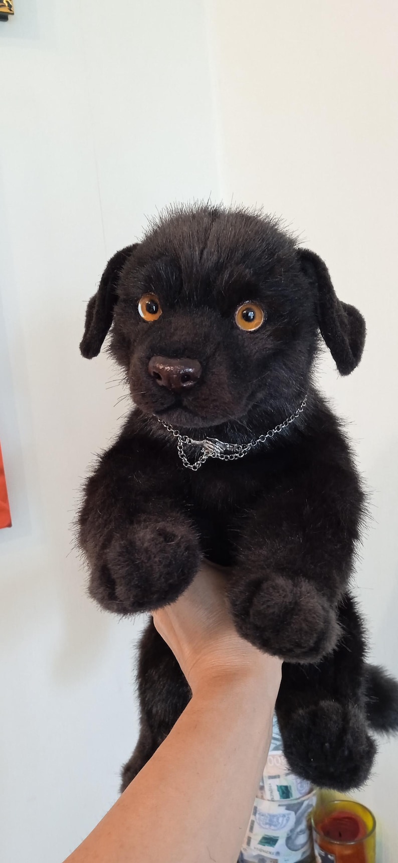 May include: A black plush puppy with amber eyes and a silver chain collar. The soft toy has dark fur and floppy ears. The puppy is being held up by a person.