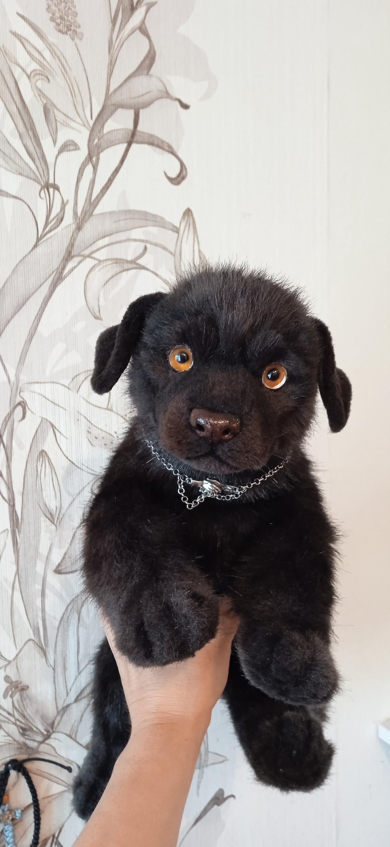 May include: A black plush puppy with amber eyes and a silver chain collar. The handmade stuffed animal has a soft, furry texture. It is held up in front of a floral patterned background.