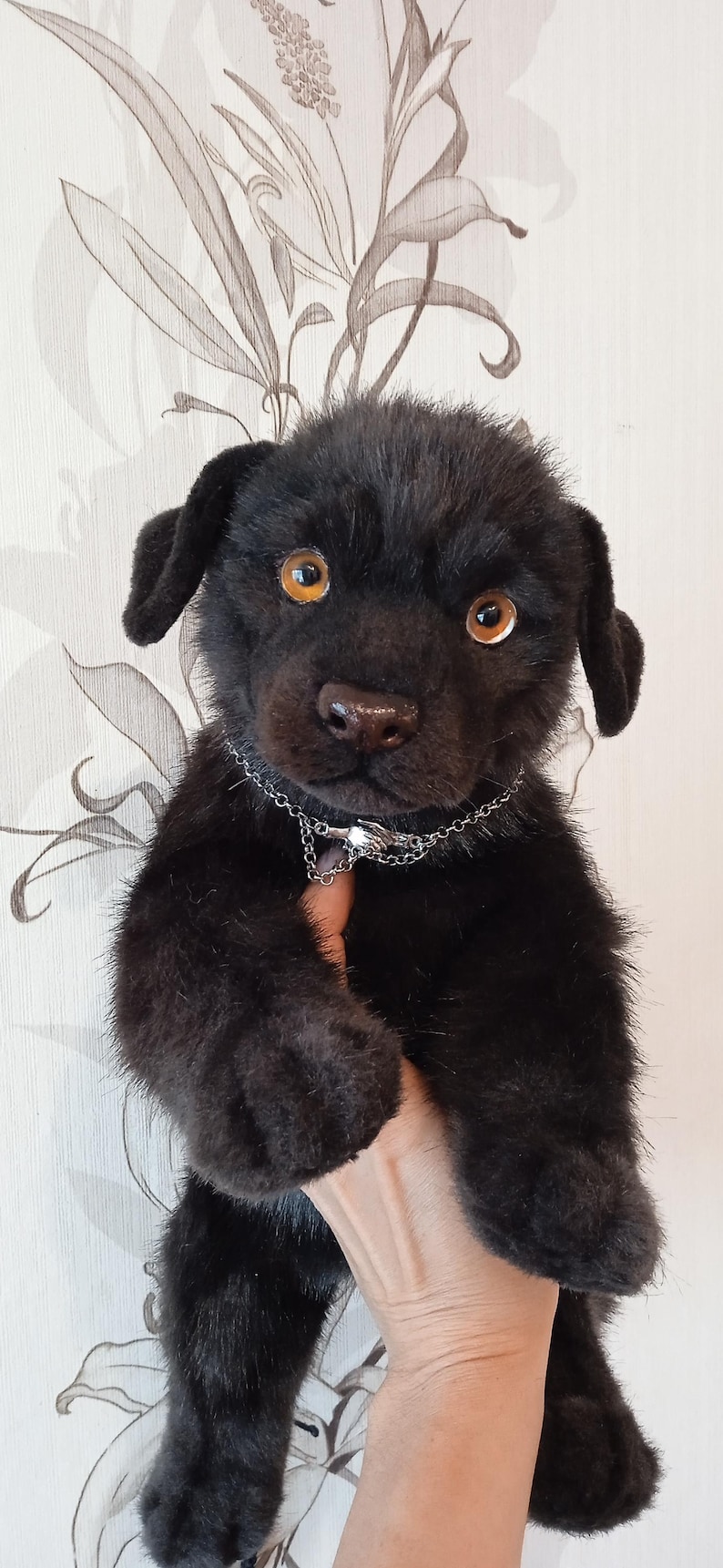 May include: A black plush puppy with amber eyes and a silver chain collar. The handmade stuffed animal has a realistic appearance and is held up against a floral patterned background.