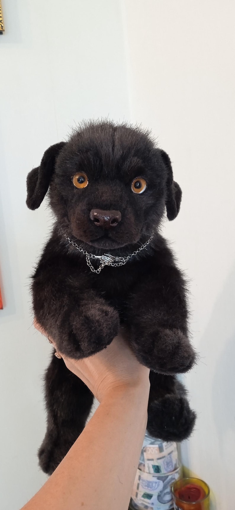 May include: A black plush puppy with amber eyes and a silver chain collar. The stuffed animal has soft, dark fur and floppy ears. The puppy is a cute, cuddly toy.