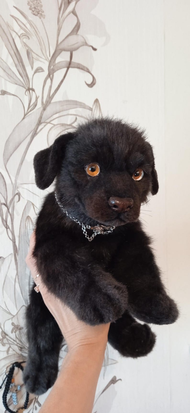May include: A black plush puppy with amber eyes and a silver chain collar. The soft toy has a realistic appearance, with soft fur and detailed features. The puppy is held up against a floral patterned background.