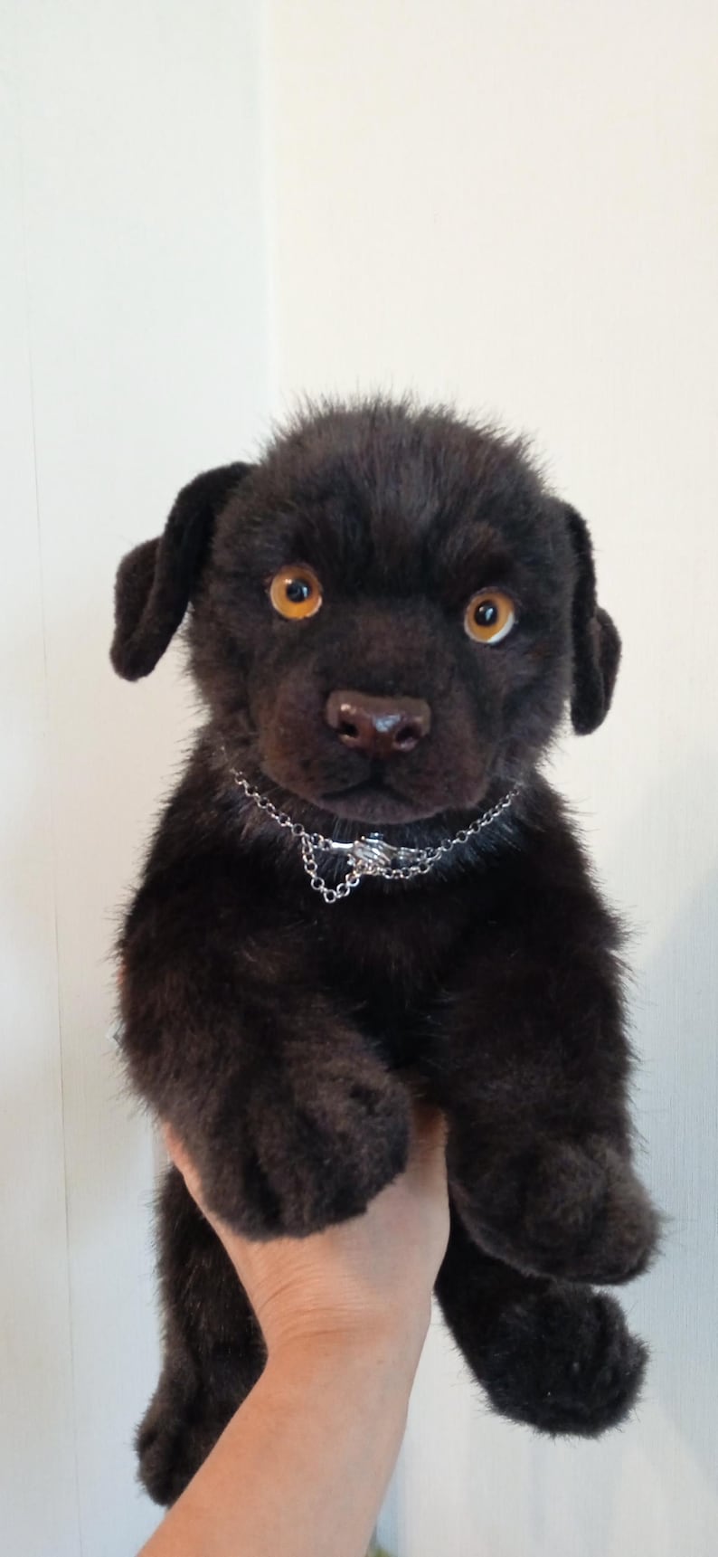 May include: A black plush puppy with amber eyes and a silver chain collar. The stuffed animal has a realistic appearance, with detailed fur and a lifelike expression. The puppy is being held up.