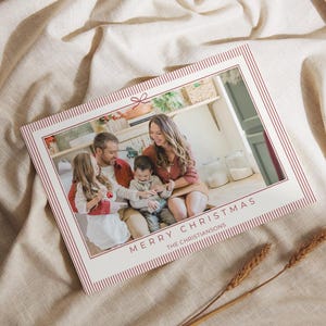 Pode incluir: Um cartão de Natal com uma foto de família no interior, com uma borda listrada vermelha e branca. O texto "MERRY CHRISTMAS" e "THE CHRISTIANSONS" é impresso em uma fonte serif. O cartão está sobre um tecido bege.