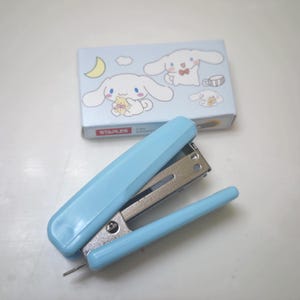 May include: A light blue stapler with a silver metal mechanism, next to a light blue box of staples. The box features illustrations of a white cartoon character with long ears. The word "STAPLES" is printed on the box.