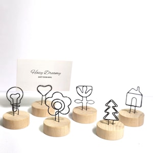 May include: A collection of decorative wire memo holders in various shapes, including a lightbulb, heart, flower, tulip, tree, and house. Each wire shape is attached to a small, round wooden base. A white card with the text "Hazy Dreamy" is also present.