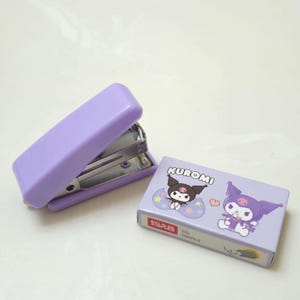 May include: A light purple stapler next to a box of staples. The box features the word "KUROMI" and illustrations of a cartoon character. The box also has the word "STAPLES" and the text "10+ 500PCS".