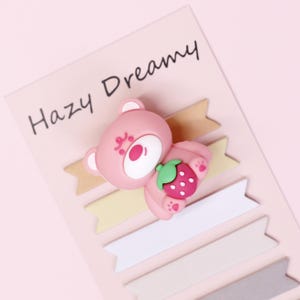 May include: A pink sticky note set featuring a pink bear holding a strawberry. The bear has a white face and ears, and the strawberry is red with green leaves. The words "Hazy Dreamy" are printed on the pink background.